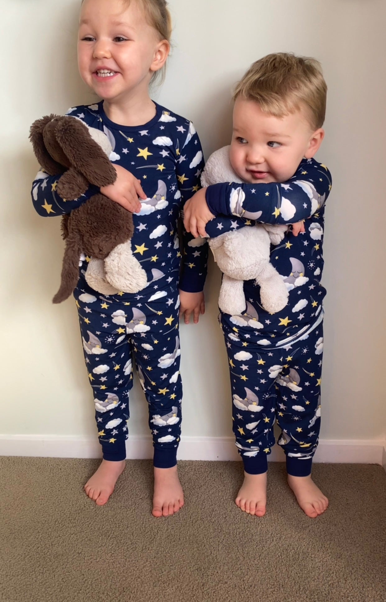 Two children wearing matching pajamas with space-themed design, holding stuffed animals against a plain background